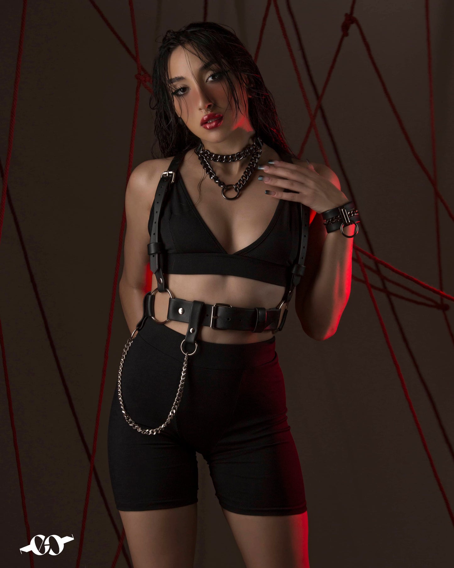A person wearing a black harness belt with a chain on the side, styled in a fashion-forward manner against a dark background with red lighting.