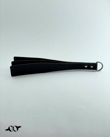 A black leather paddle with a ring at the handle, laid out flat on a white background.