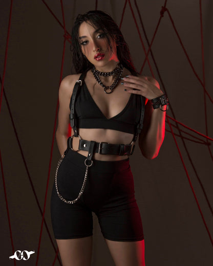 A person wearing a black harness belt with a chain on the side, styled in a fashion-forward manner against a dark background with red lighting.