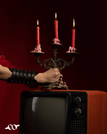 A person's wrist wearing a wide leather bracelet with multiple straps and studs, displayed in front of a red-lit candelabra on top of an orange television set.
