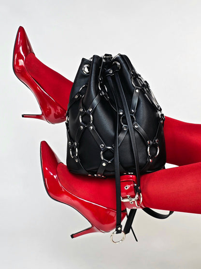 A black leather bucket bag with silver-tone chain and stud embellishments, styled between a model's legs wearing bright red patent leather high heels and red tights against a light gray background.