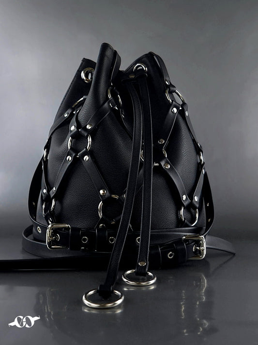 A black leather bucket bag with an intricate strap and buckle harness design, featuring silver-tone metal hardware and a drawstring closure, set against a dark gray reflective background.